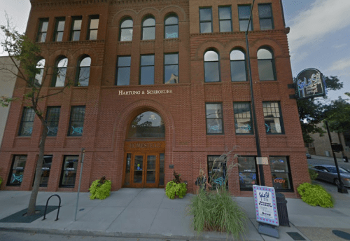 Historic brick building with "Hartung & Schroeder" sign, featuring large windows and a neon sign, located on a city street.