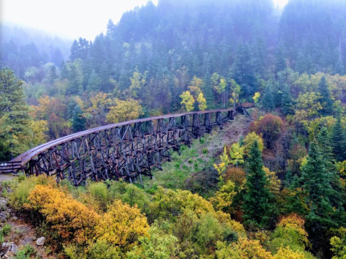 A wooden trestle bridge spans a valley surrounded by colorful autumn trees and misty mountains.