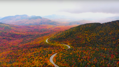 A winding road cuts through vibrant autumn foliage in a mountainous landscape, showcasing rich reds, oranges, and yellows.