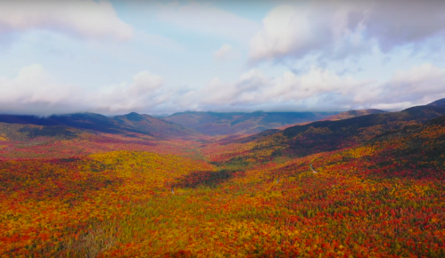 Aerial view of a vibrant autumn landscape with colorful foliage covering rolling hills and mountains under a blue sky.