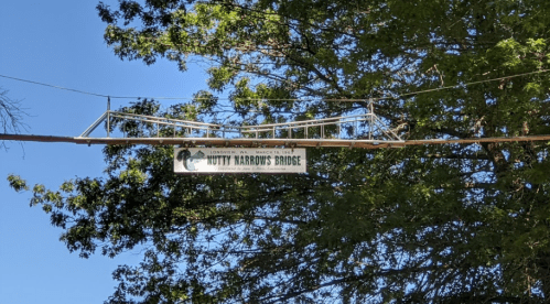 Sign for Nutty Harmon's Bridge hanging above a tree-lined area, with clear blue sky in the background.
