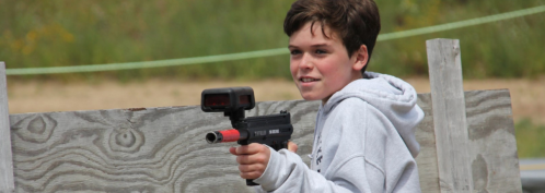 A young boy in a hoodie aims a toy gun while standing behind a wooden barrier outdoors.