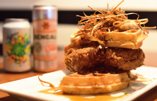 A plate featuring fried chicken atop waffles, garnished with crispy onions, alongside two cans of colorful beverages.