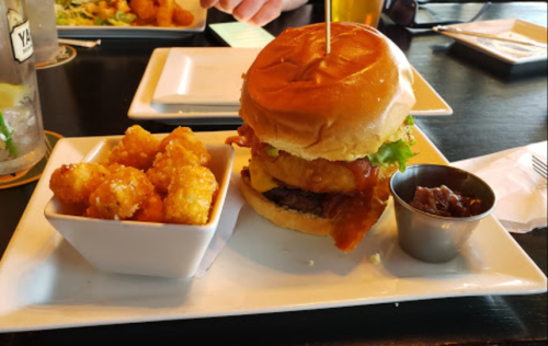 A burger with bacon, lettuce, and onion rings, served with tater tots and a small cup of sauce on a white plate.