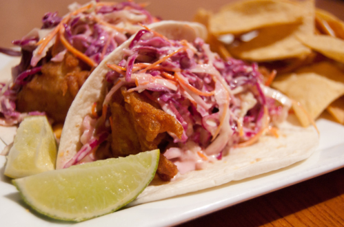 Two crispy fish tacos topped with colorful slaw, served with lime wedges and a side of tortilla chips.