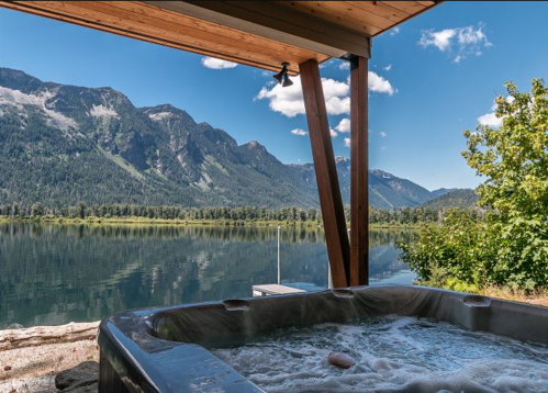 A hot tub on a deck overlooking a serene lake and mountains under a clear blue sky.