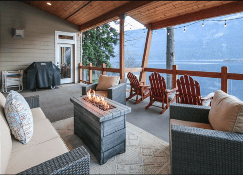 Cozy outdoor seating area with a fire pit, overlooking a scenic lake and mountains, under a wooden porch.