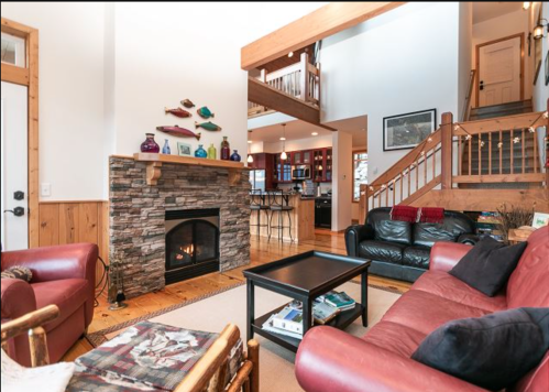Cozy living room with a stone fireplace, red leather sofas, and a view of a modern kitchen and wooden staircase.
