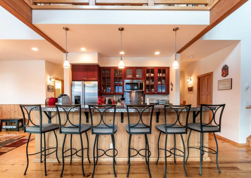 Modern kitchen with a wooden bar, high stools, stainless steel appliances, and warm lighting.