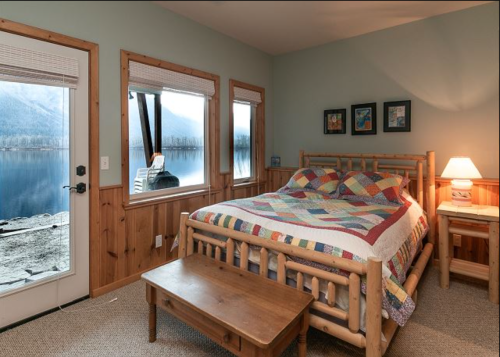 Cozy bedroom with a quilted bed, wooden furniture, and large windows overlooking a serene lake and mountains.