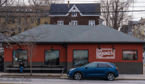 A red building with a sign reading "The Sandwich Hut" and a blue car parked outside on a street.