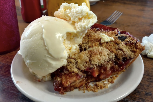 A slice of fruit pie topped with a scoop of vanilla ice cream on a white plate, with drinks in the background.
