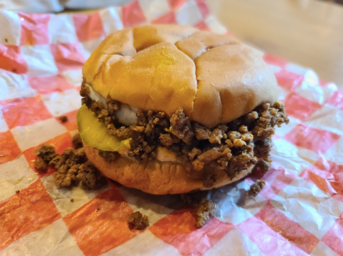 A close-up of a sandwich with a soft bun, filled with seasoned ground meat and a slice of pickle, resting on checkered paper.