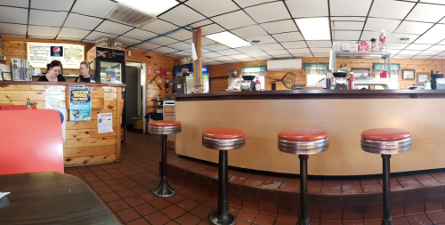 A cozy diner interior with wooden walls, a counter, and red stools. Two staff members are chatting behind the counter.