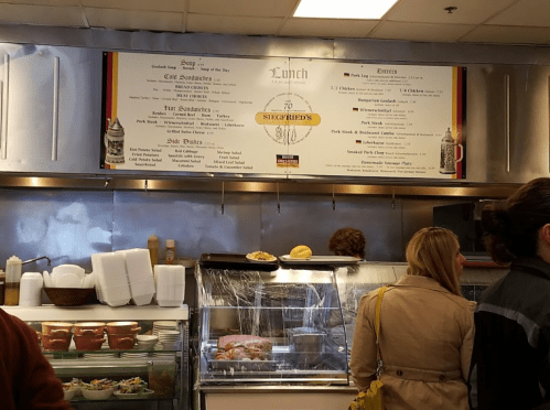 A menu board at a deli featuring lunch options, including sandwiches, soups, and salads, with patrons in the background.