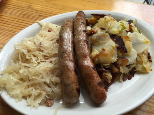 A plate with two sausages, sautéed potatoes, and a side of sauerkraut on a wooden table.