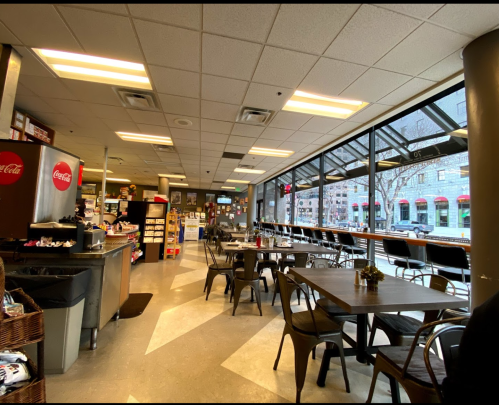 A cozy café interior with tables, chairs, and a large window overlooking the street, featuring a counter and shelves.