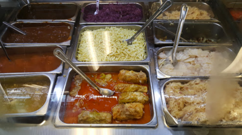 A variety of dishes in metal trays, including sauces, noodles, and stuffed rolls, displayed in a food service setting.
