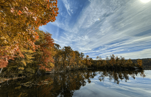 A serene lake surrounded by autumn trees, reflecting colorful foliage and a blue sky with wispy clouds.