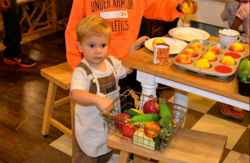 A young child in overalls holds a basket of colorful toy fruits in a playful kitchen setting.