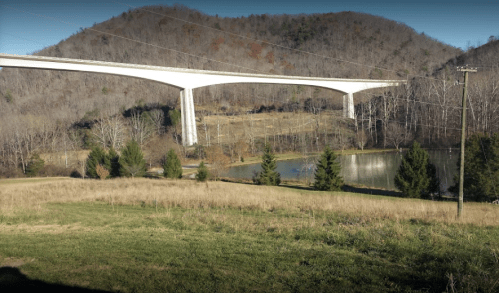 A large white bridge spans over a calm pond, surrounded by trees and rolling hills in a rural landscape.