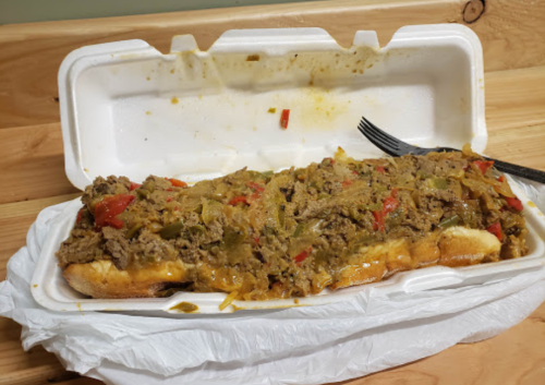 A large sandwich filled with seasoned beef, peppers, and onions, served in a takeout container with a fork.