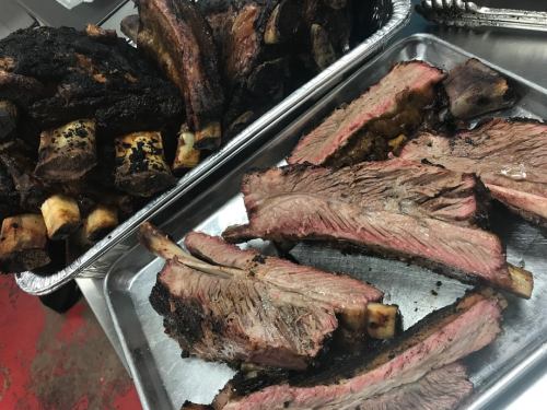 Two trays of smoked beef ribs, one with whole ribs and the other with sliced pieces, showcasing a smoky, charred exterior.