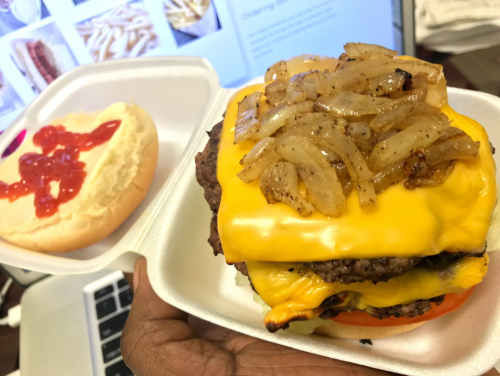 A hand holding a cheeseburger with grilled onions, tomato, and a bun, alongside a plain bun with ketchup.