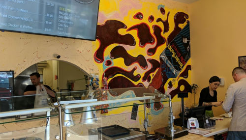 A colorful mural on a restaurant wall with a counter where a customer is being served by a staff member.