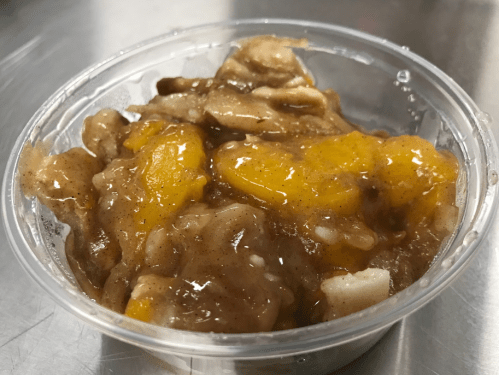A clear cup filled with a warm fruit mixture, featuring chunks of peaches and a cinnamon glaze.