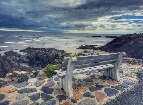 A wooden bench overlooks a rocky beach under a cloudy sky, with gentle waves lapping at the shore.
