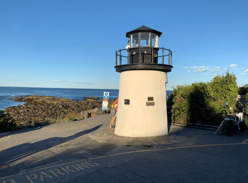 A small lighthouse stands by the shore, with rocky coastline and blue sky in the background.