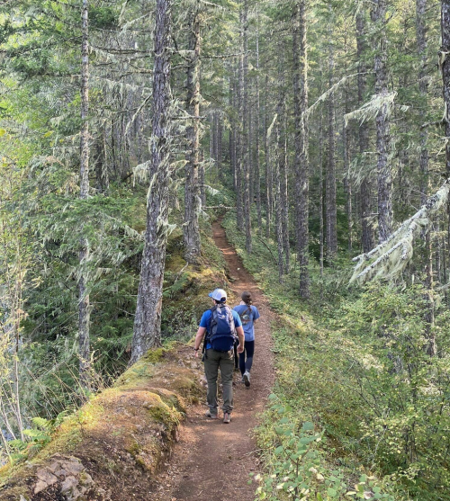 Two hikers walking along a narrow trail through a lush, green forest with tall trees.