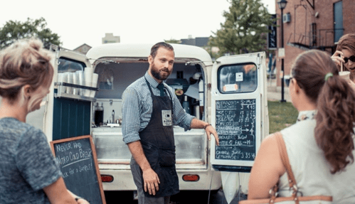 A barista serves customers at a food truck, displaying a menu on a chalkboard in an outdoor setting.