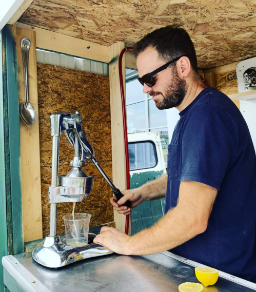 A man in sunglasses uses a manual juicer to squeeze juice into a glass, surrounded by a rustic wooden backdrop.