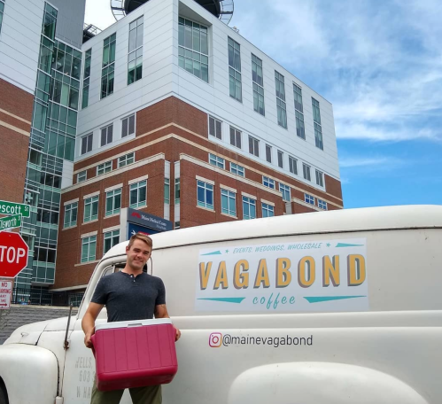 A man stands next to a coffee van with a cooler, in front of a modern building. The van is branded "Vagabond Coffee."