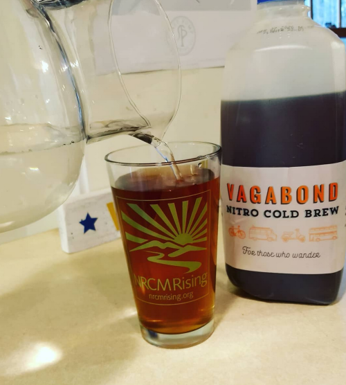 A glass with a logo is filled with dark liquid from a bottle labeled "Vagabond Nitro Cold Brew."