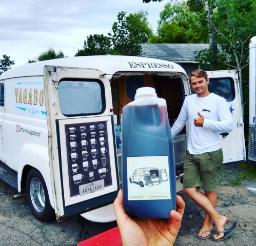 A person stands by a vintage coffee truck, holding a large bottle of coffee, with a thumbs-up gesture.