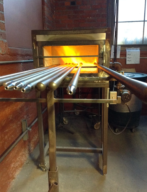 A glassblowing furnace with glowing interior and metal rods extending from it, set in a workshop environment.