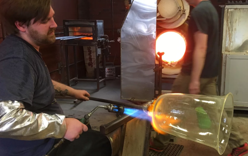 A glassblower shapes molten glass with a torch in a workshop, with glowing furnaces in the background.