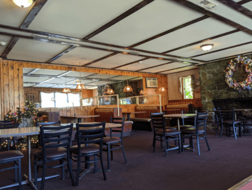 Cozy restaurant interior with wooden walls, tables, and chairs, featuring a stone fireplace and decorative wreath.