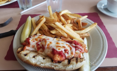 A plate with a breaded chicken sandwich topped with marinara and cheese, served with fries and pickles.