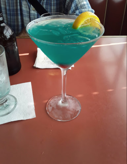 A blue cocktail in a large glass garnished with a lemon slice, sitting on a pink table.