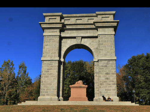 A large stone archway with a lion statue at its base, surrounded by trees and a clear blue sky.