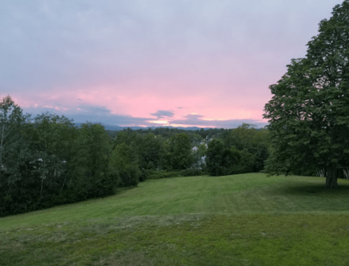 A serene landscape at sunset, featuring a grassy field, trees, and a colorful sky with pink and purple hues.