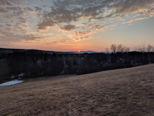 A serene sunset over a landscape with rolling hills, trees, and scattered clouds in a colorful sky.