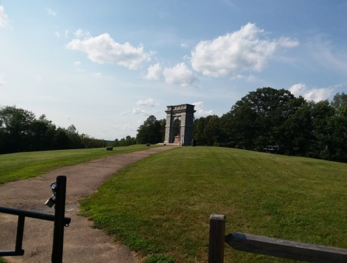 A stone monument on a grassy hill under a blue sky with clouds, surrounded by trees.