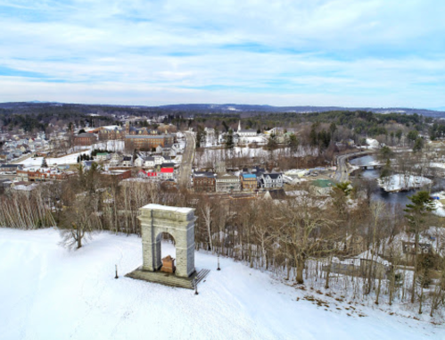 Aerial view of a snowy landscape featuring a monument, town buildings, and a river in the background.