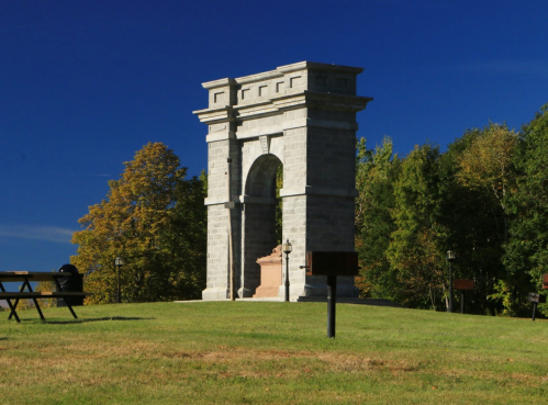 A stone arch monument on a grassy hill, surrounded by trees under a clear blue sky.