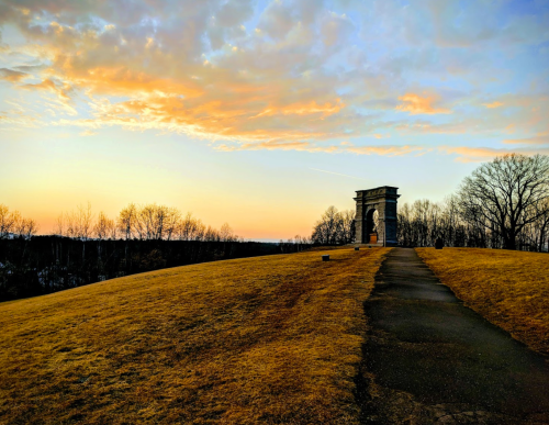 A stone archway on a grassy hill at sunset, with colorful clouds and bare trees in the background.
